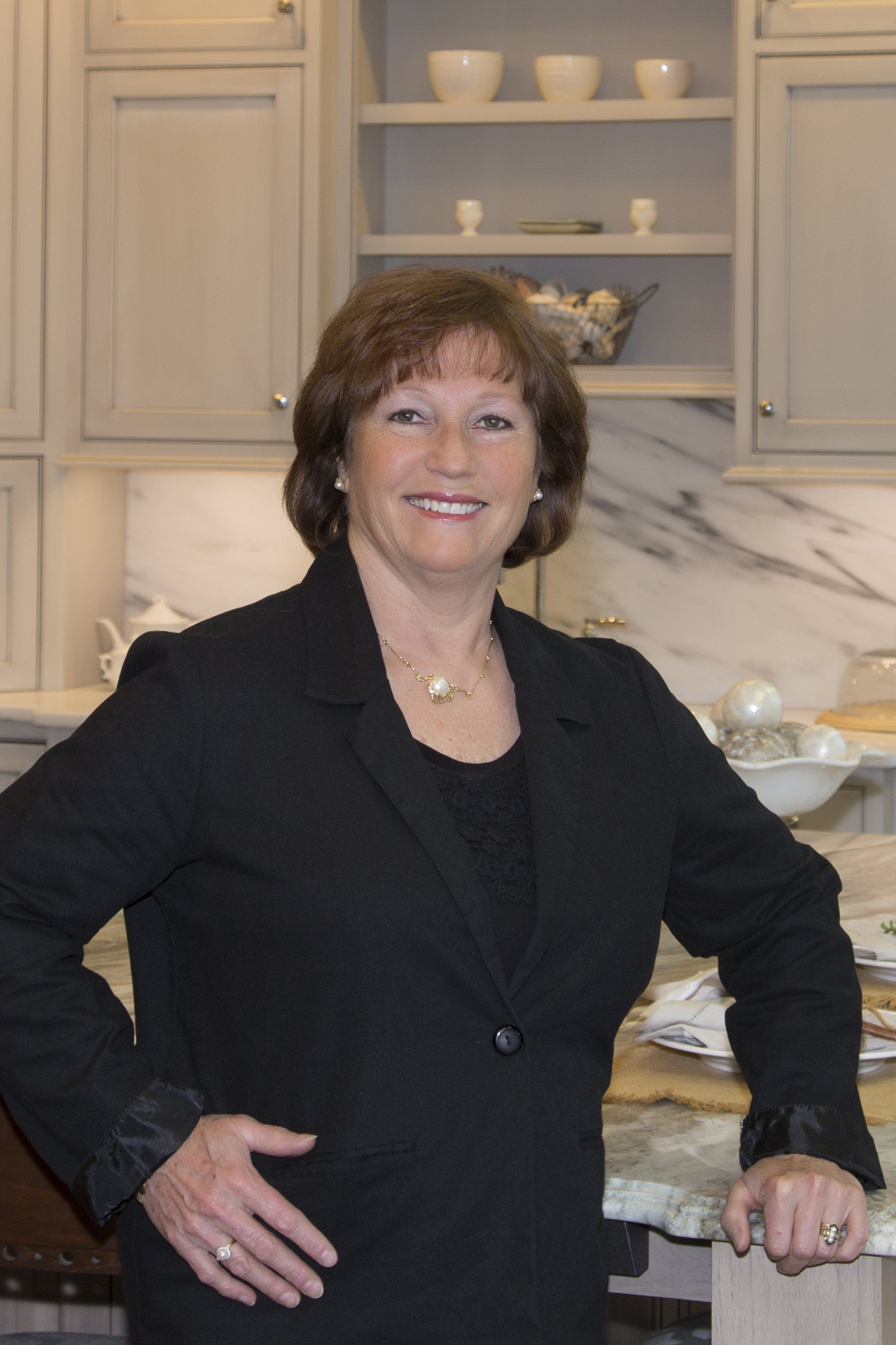 Woman in a black blazer smiling in a bright kitchen with marble countertops and light cabinetry