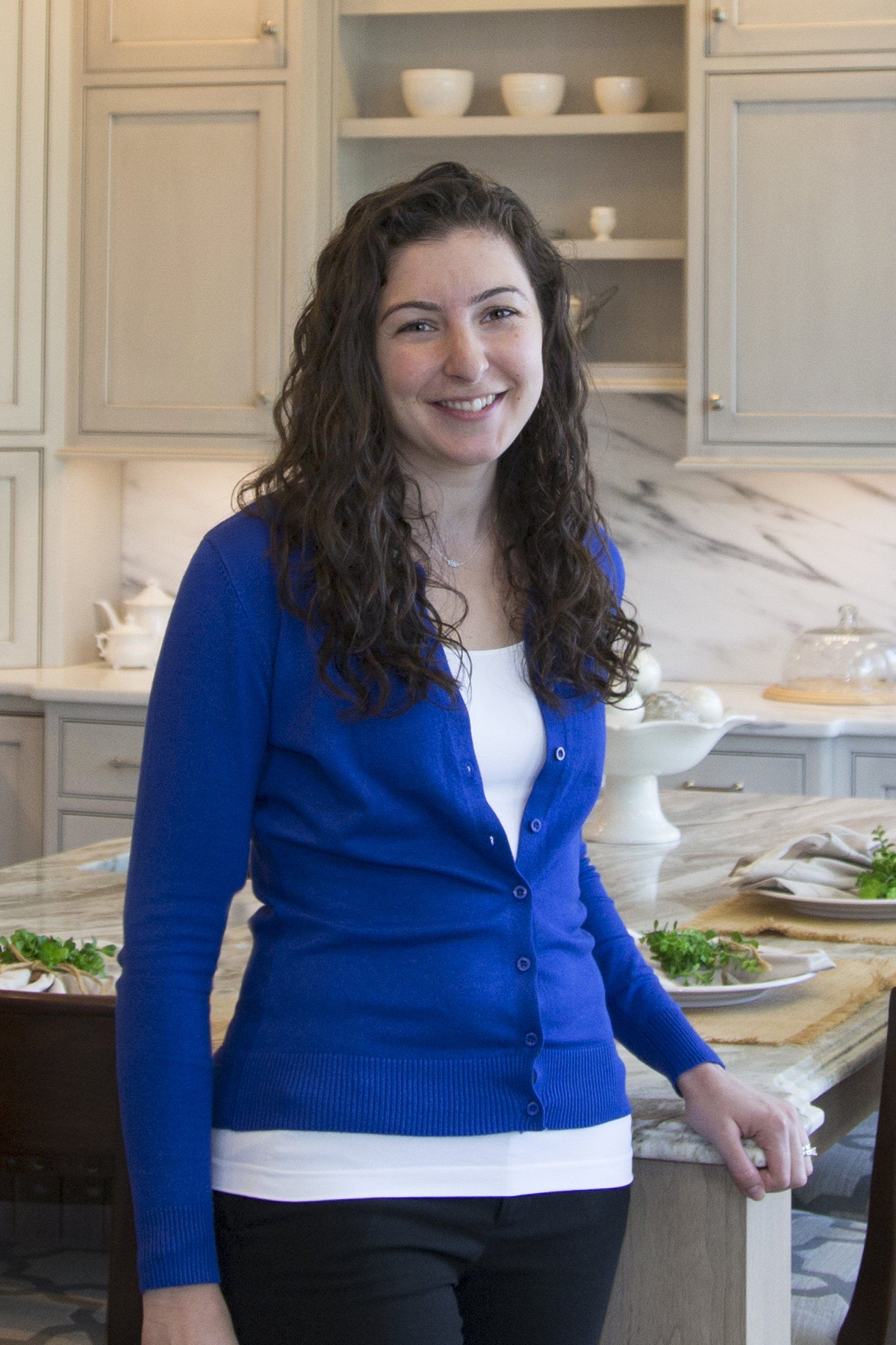 Woman in a blue cardigan standing in a bright kitchen, smiling beside a counter with food.