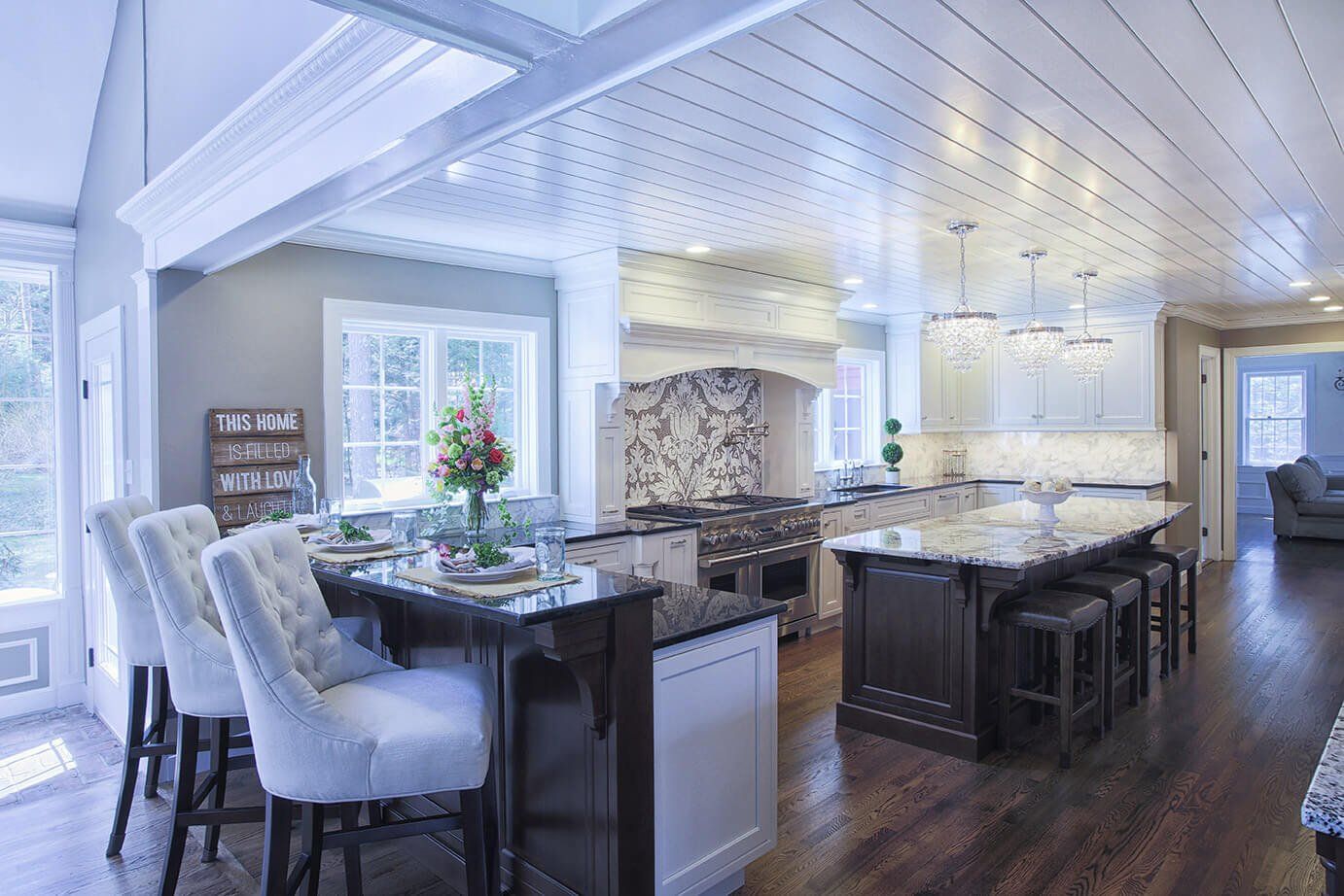 Bright open-concept kitchen and dining area with white beams, dark island stools, and light blue upholstered chairs