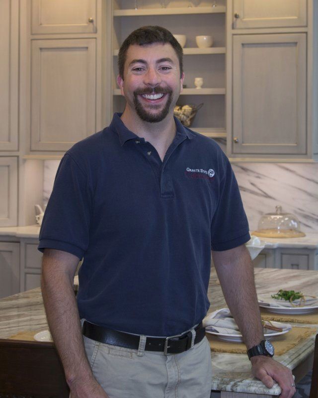 Smiling man in navy polo and khaki pants standing in a bright kitchen with a marble island