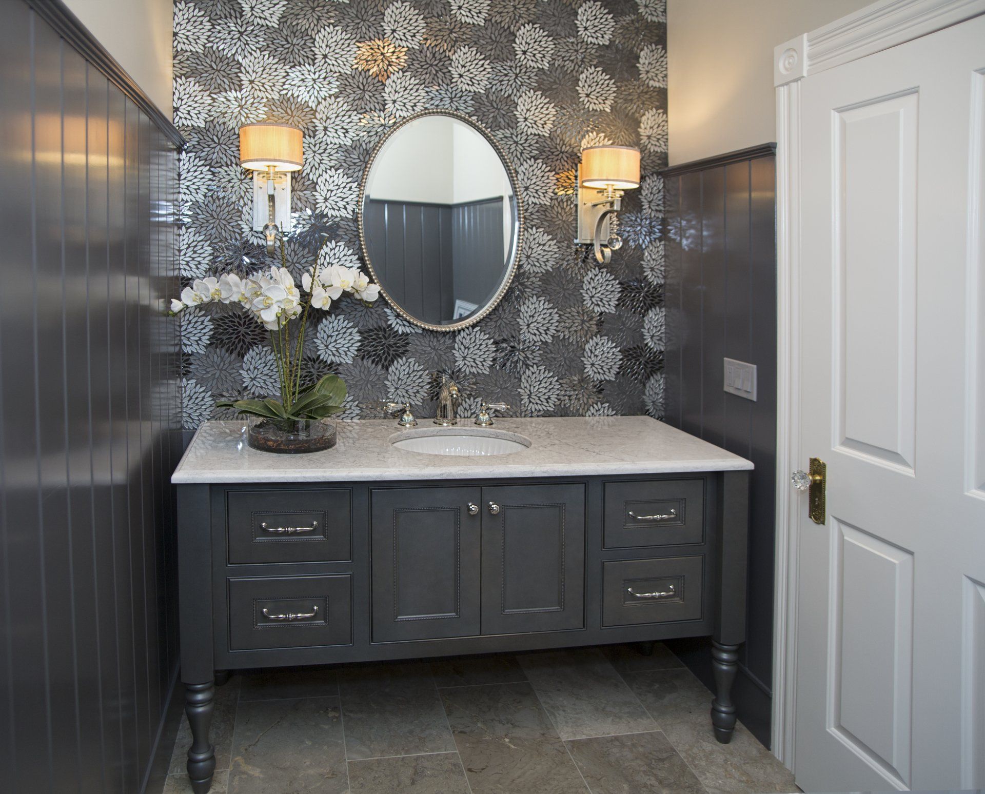 Elegant dark-gray bathroom vanity with marble top, round mirror, floral wallpaper, and wall sconces