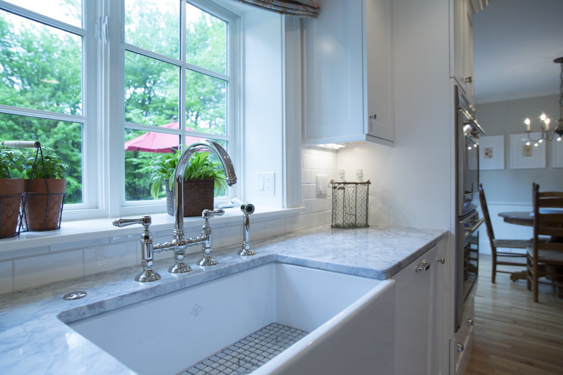 Bright kitchen sink beneath large window, with marble countertop and white cabinetry