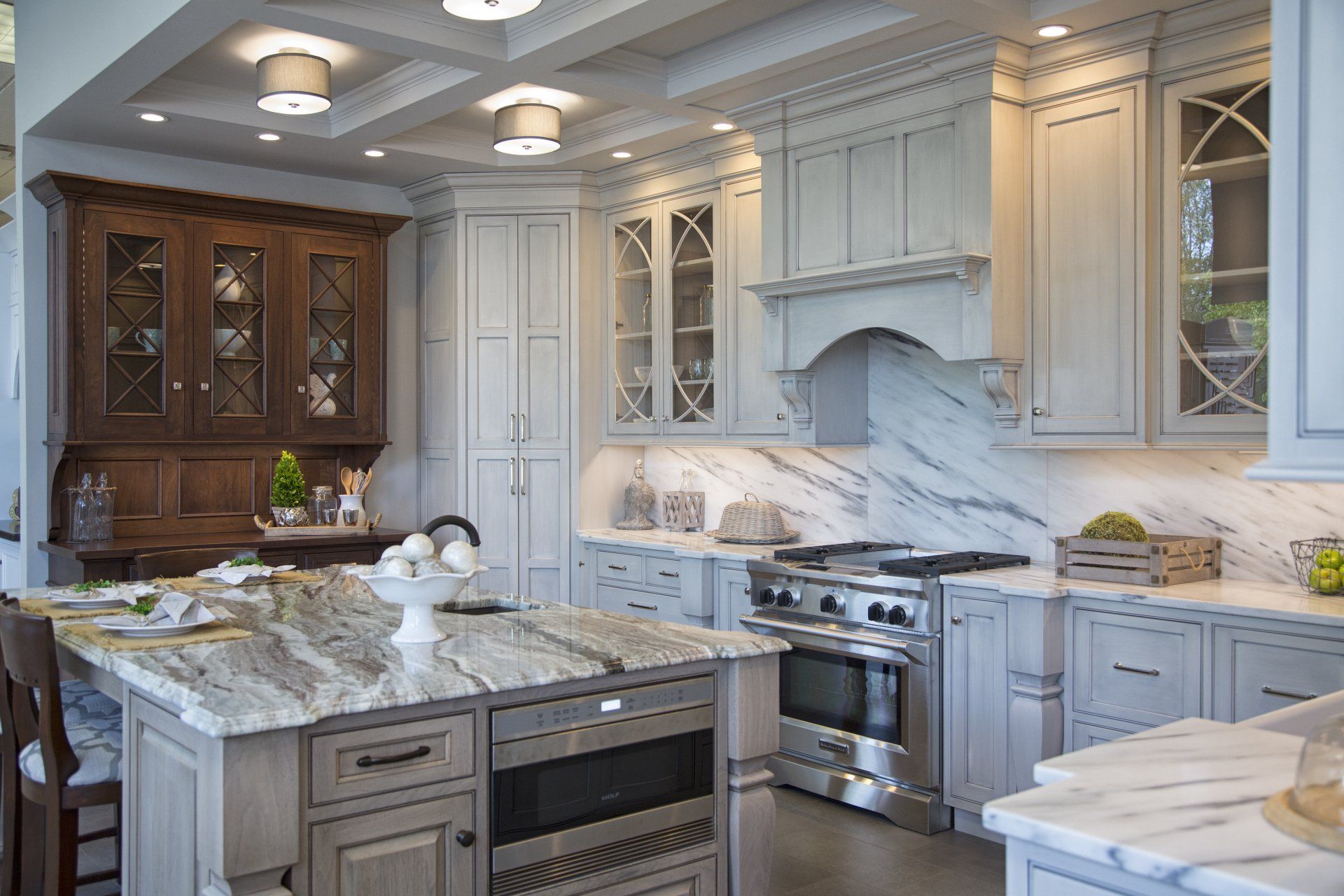Bright white kitchen with marble island, gray cabinets, stainless stove, and glass-front display cabinets