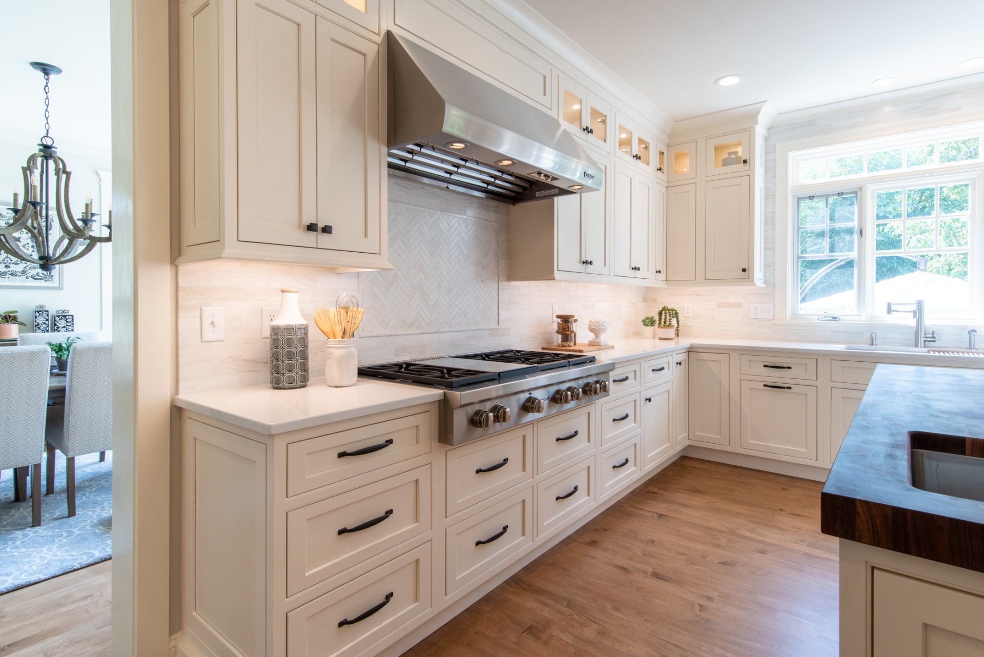 Bright white kitchen with farmhouse cabinets, stainless range hood, wood floor, and a sunlit window above the sink