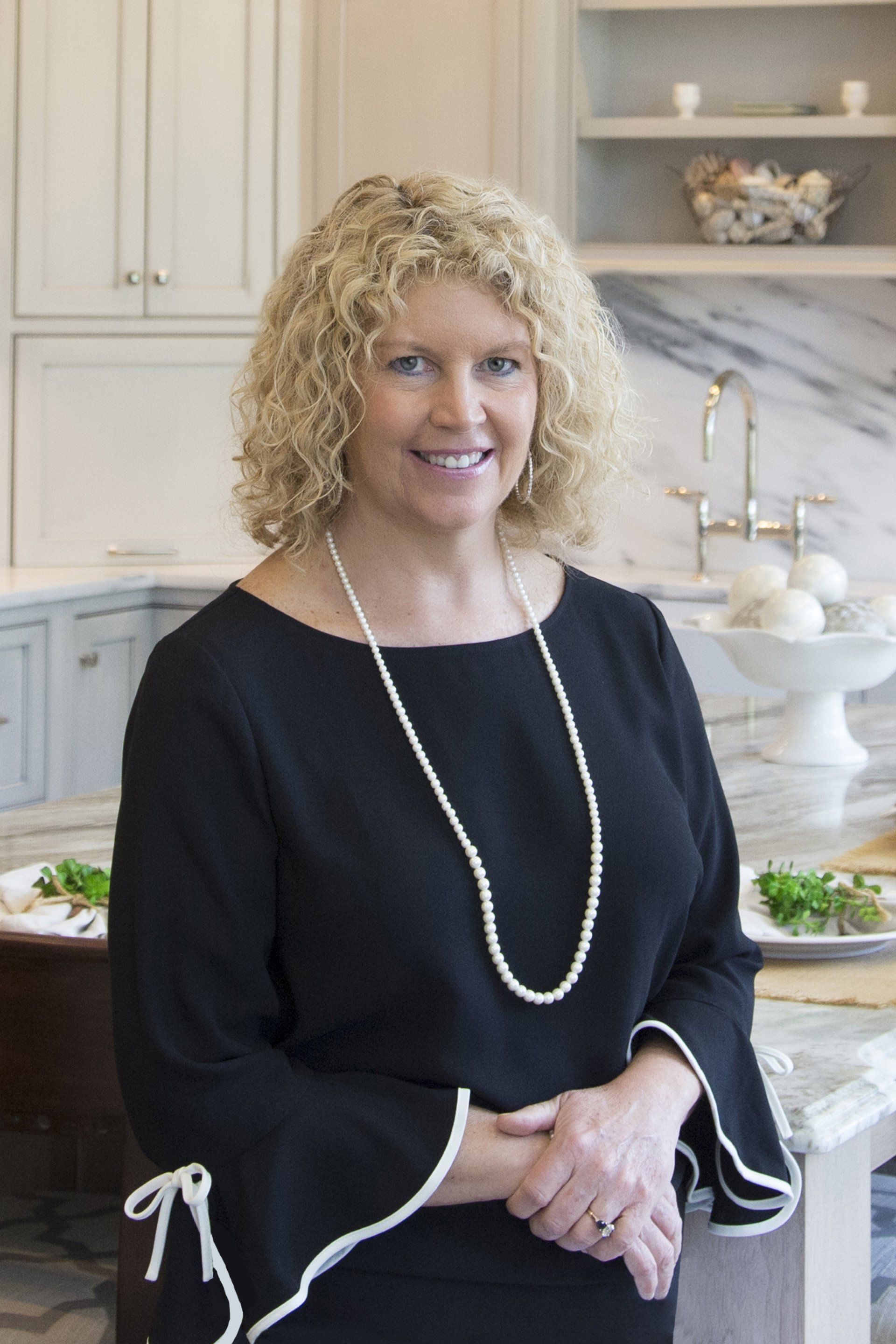 Woman standing in a bright kitchen, wearing a black top and pearl necklace, smiling at the camera