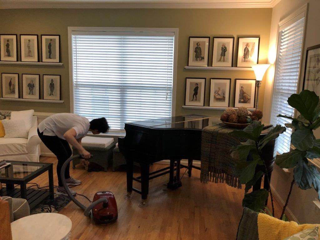 Person cleaning a piano in a sunlit living room with framed art and plants.