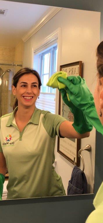 Woman in a green shirt smiling while cleaning a bathroom mirror with a green cloth.