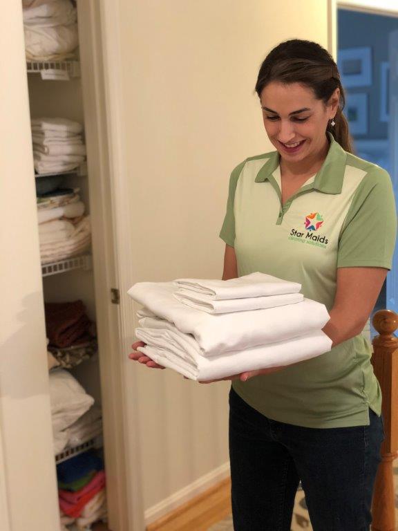 Woman in green polo carrying folded towels in a hallway beside an open linen closet