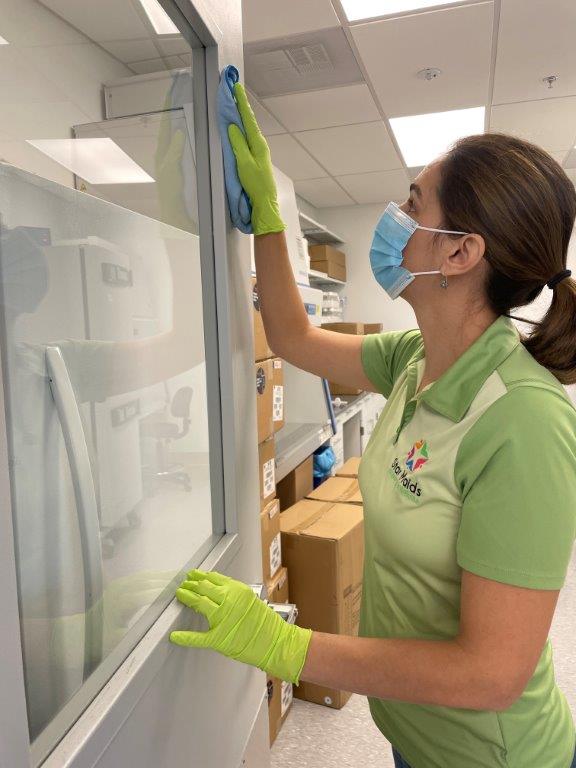 Woman in green polo and gloves cleaning a glass wall in an office hallway.