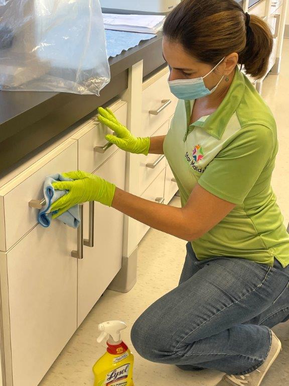 Person in green gloves disinfects a cabinet handle in a lab, with cleaning spray nearby.