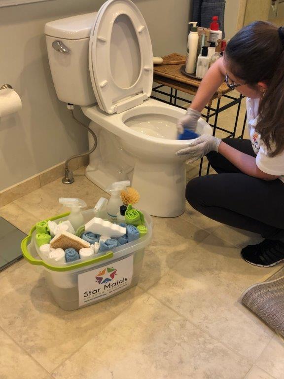 Person cleaning a toilet with gloves beside a bin of cleaning supplies in a bathroom.