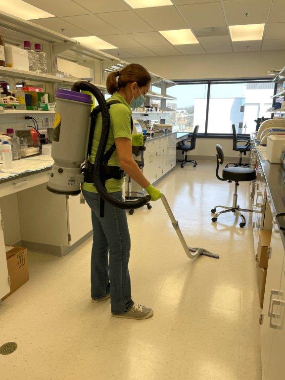 Person using a backpack vacuum cleans a bright lab floor with a hose wand.