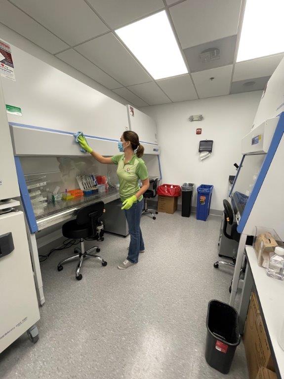 Person cleaning a lab bench in a small laboratory, wearing a green shirt and jeans.