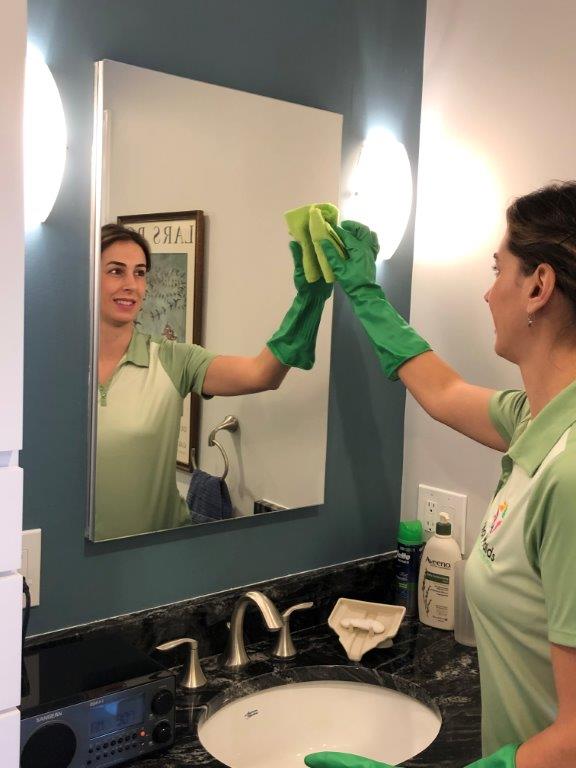 Person cleaning a bathroom mirror with a green cloth, reflected above a sink and countertop.