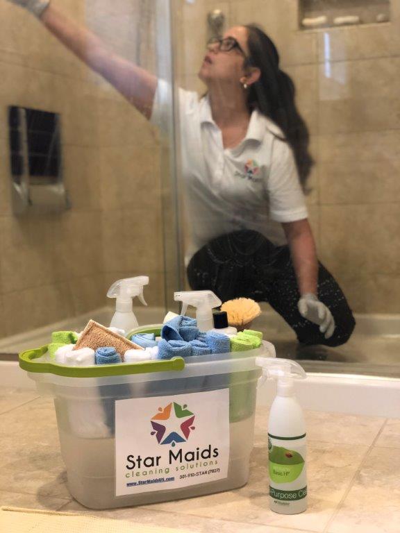 Cleaner kneeling in a bathroom, reaching up to wipe a shower wall beside a Star Maids cleaning caddy.