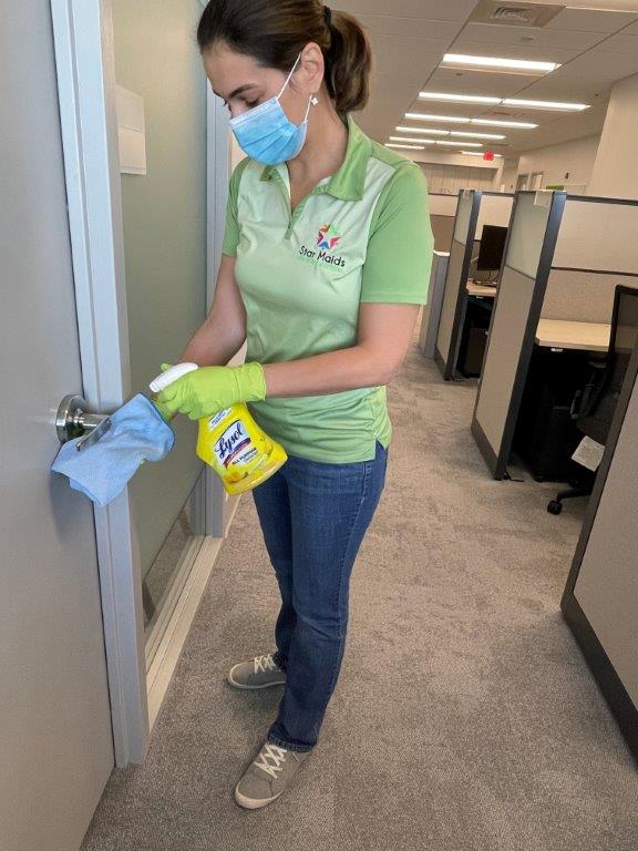 Person in green shirt and mask disinfecting a door handle in an office hallway