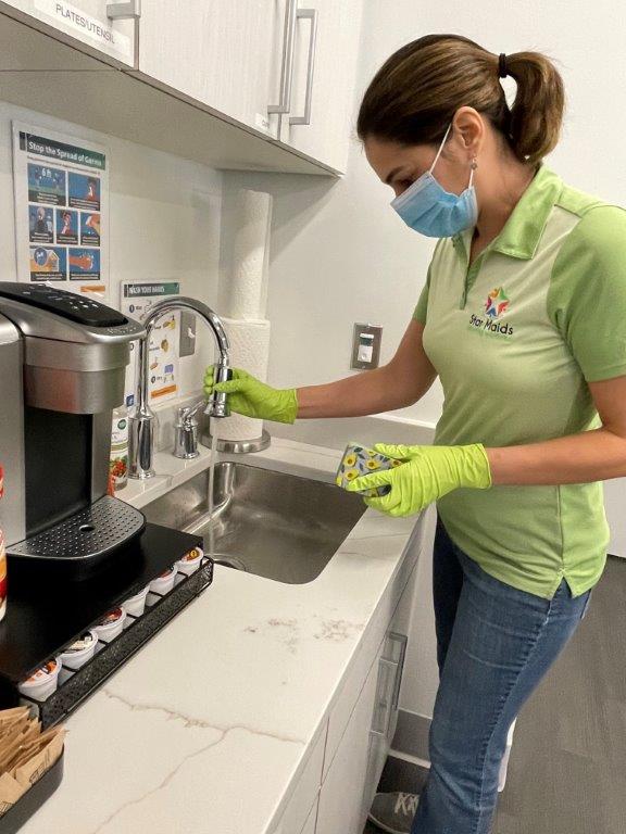 Woman in green shirt washing hands at a sink beside a coffee machine in a kitchen break area