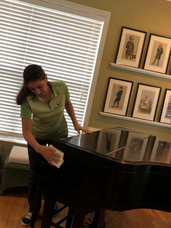 Woman wiping a black grand piano in a bright room with framed art on the wall.