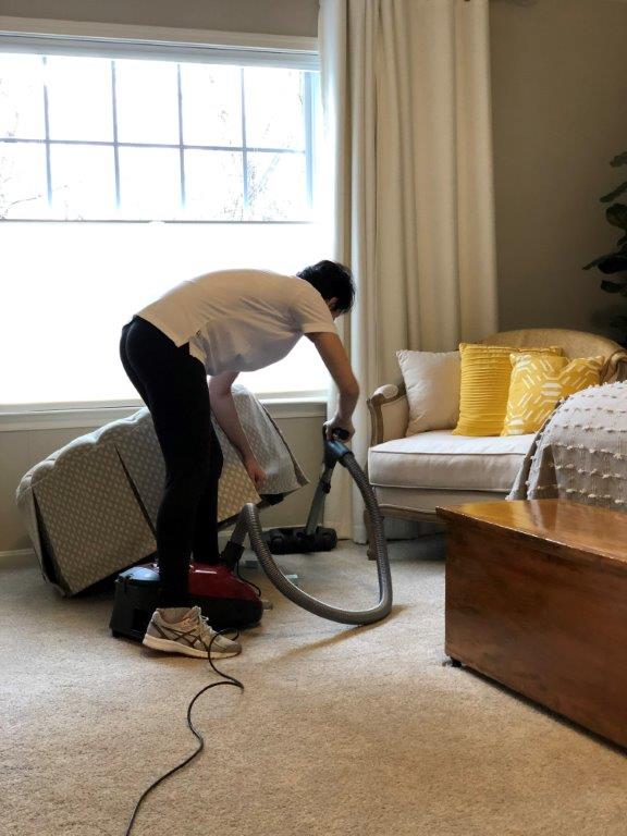 Person vacuuming a carpet in a bright living room beside a sofa and window