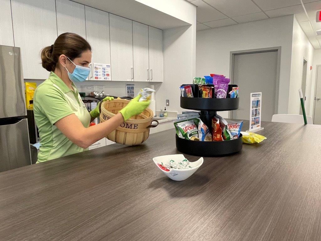 Woman in green scrubs sorting snacks in a break room next to a table with a snack carousel.