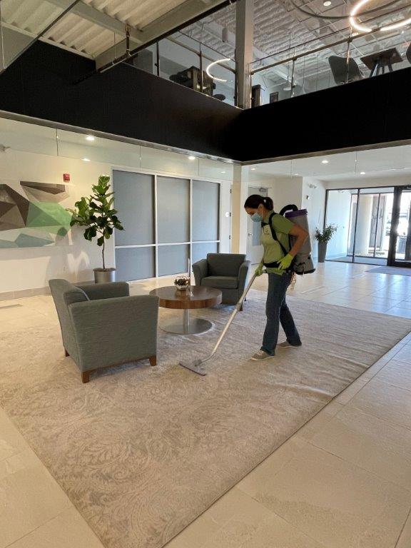 Person vacuuming a beige rug in a modern office lounge with gray chairs and a coffee table