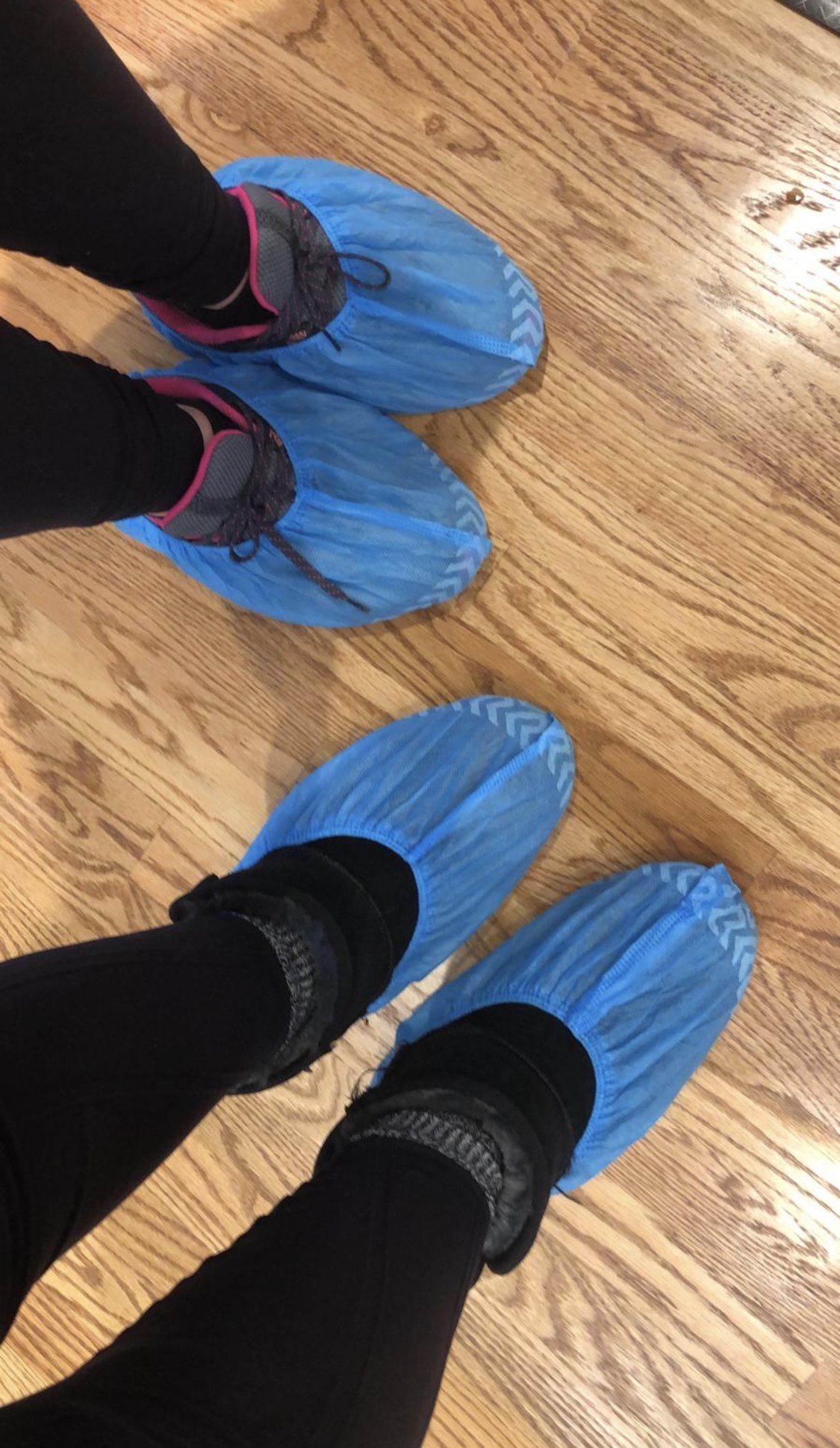 Three pairs of feet wearing blue disposable shoe covers on a hardwood floor