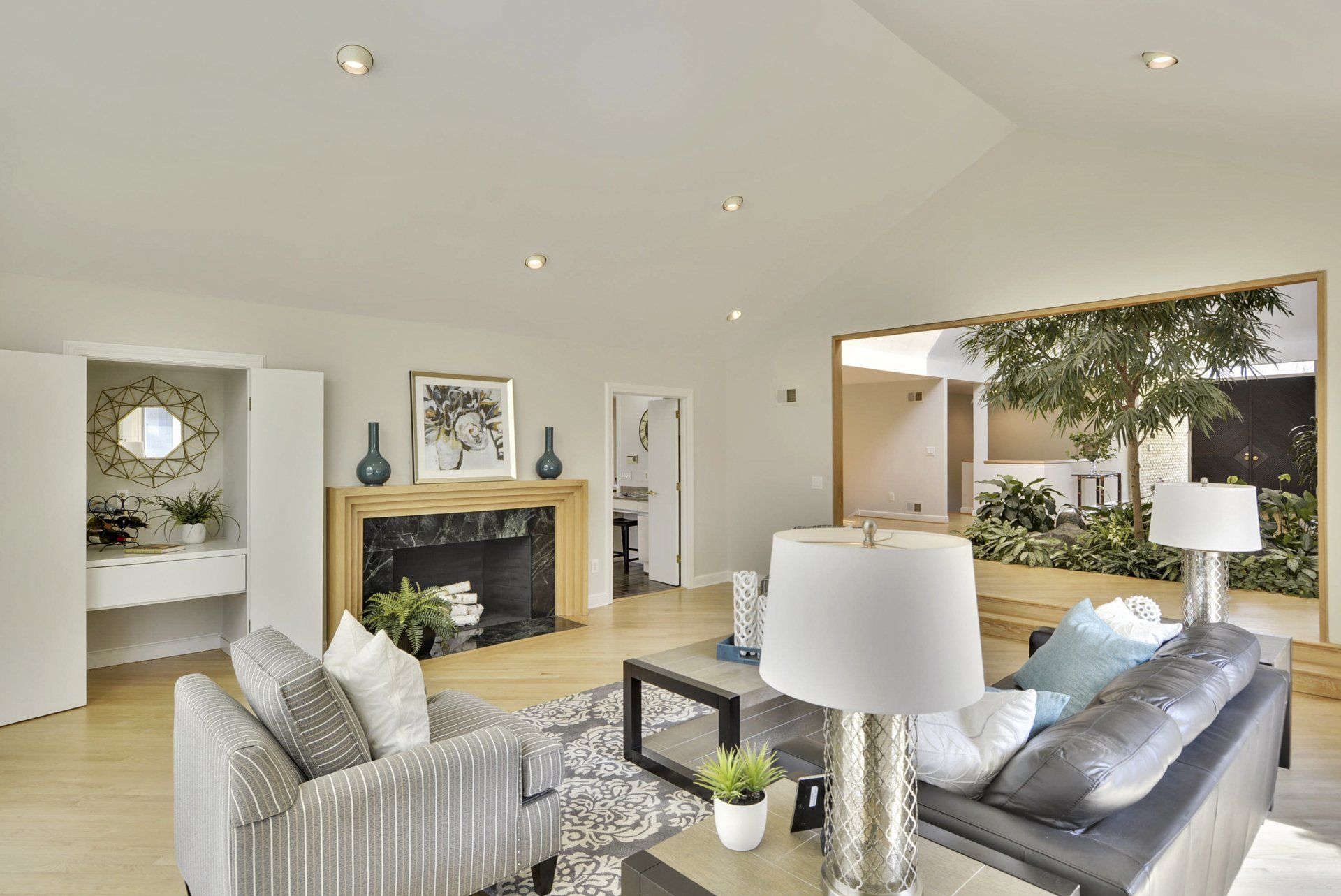 Bright living room with fireplace, striped chairs, dark sofa, and open view to a patio with tree and table lamp.