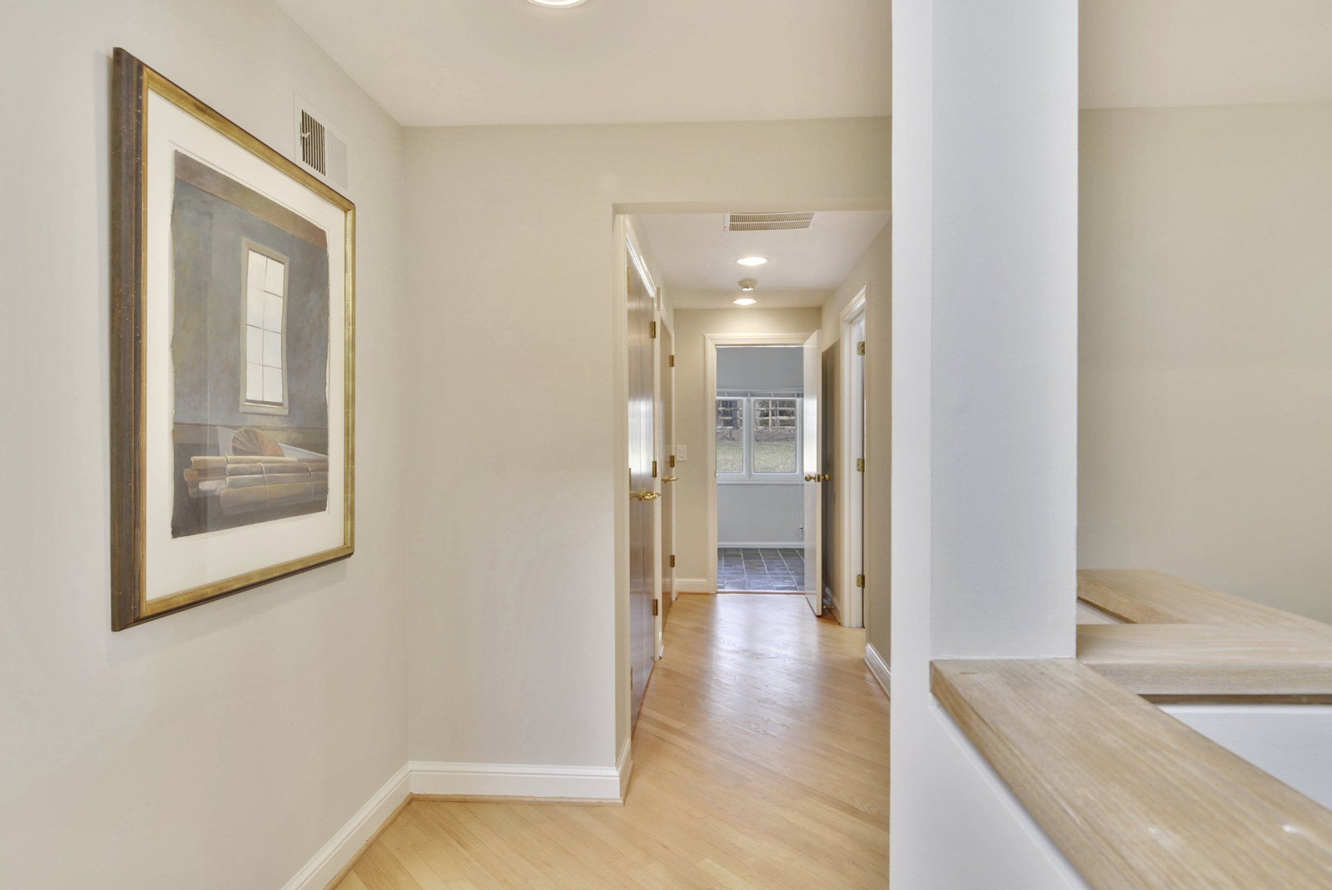 Bright hallway with hardwood floors, framed wall art, and a view into a light-filled room ahead.