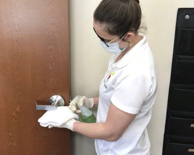 Woman in white uniform disinfecting a door handle with a spray bottle and cloth.
