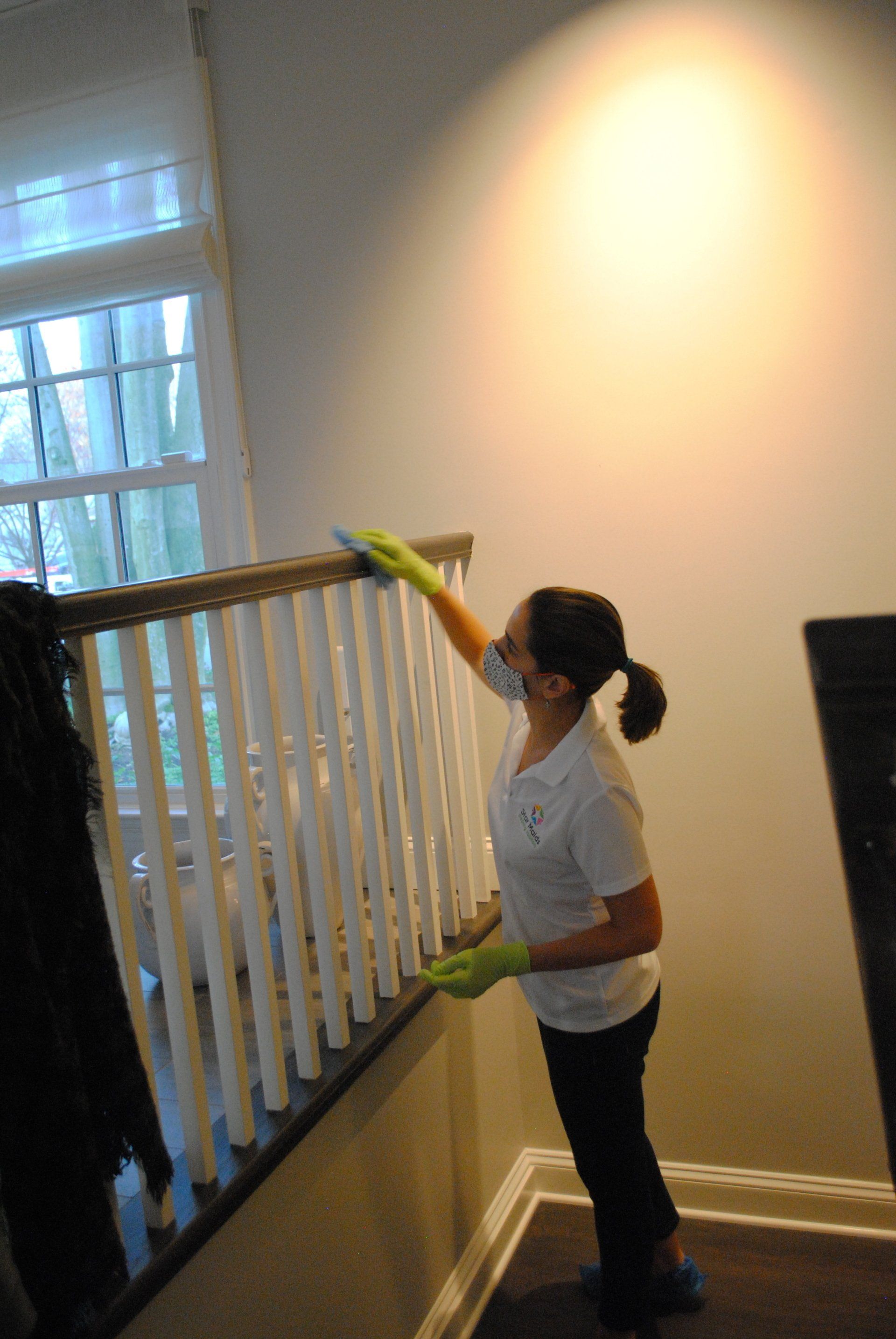 Person cleaning a stair railing beside a window in a hallway, using a green cloth or glove