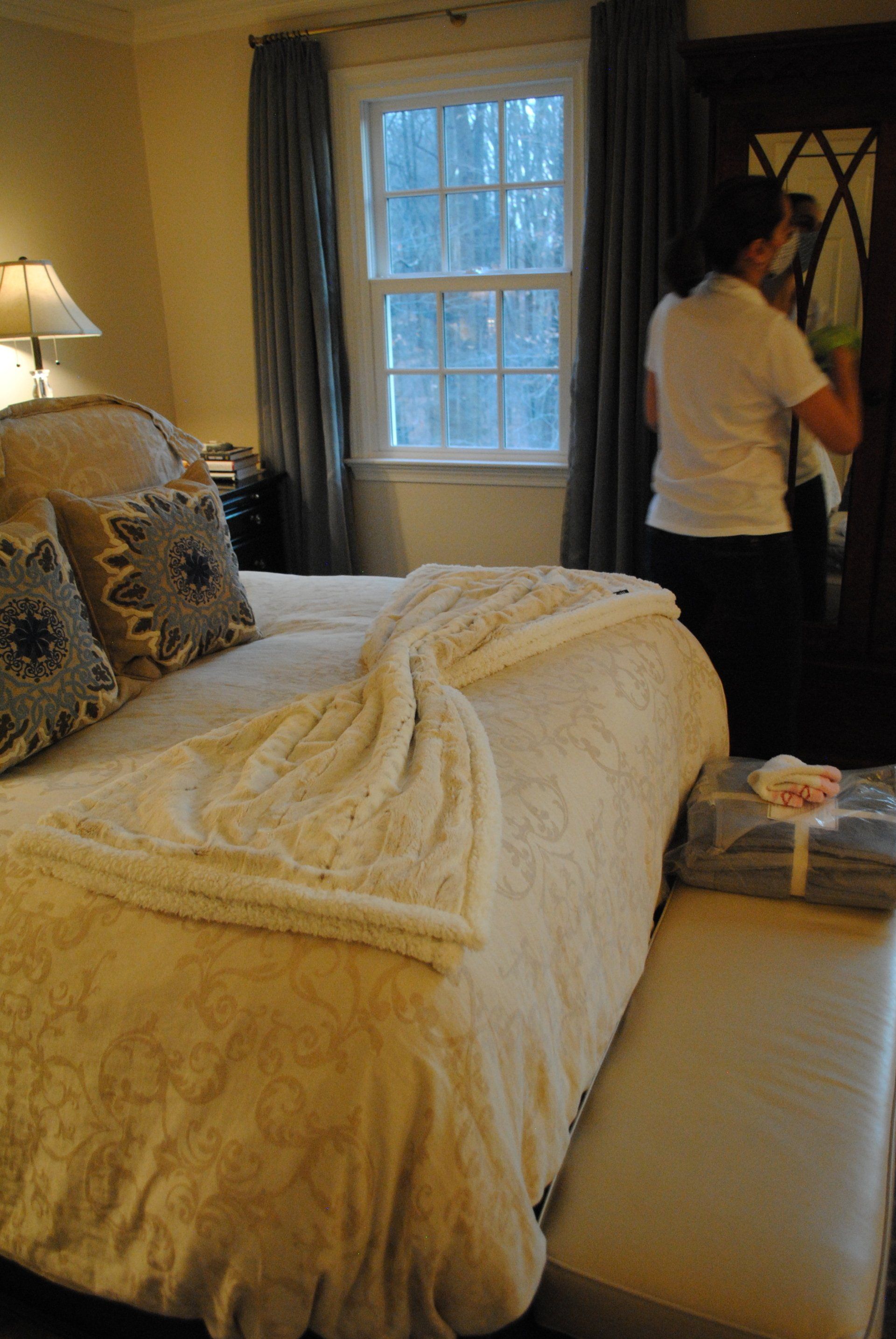 Bedroom with a made bed, window, lamp, and a person standing by the door