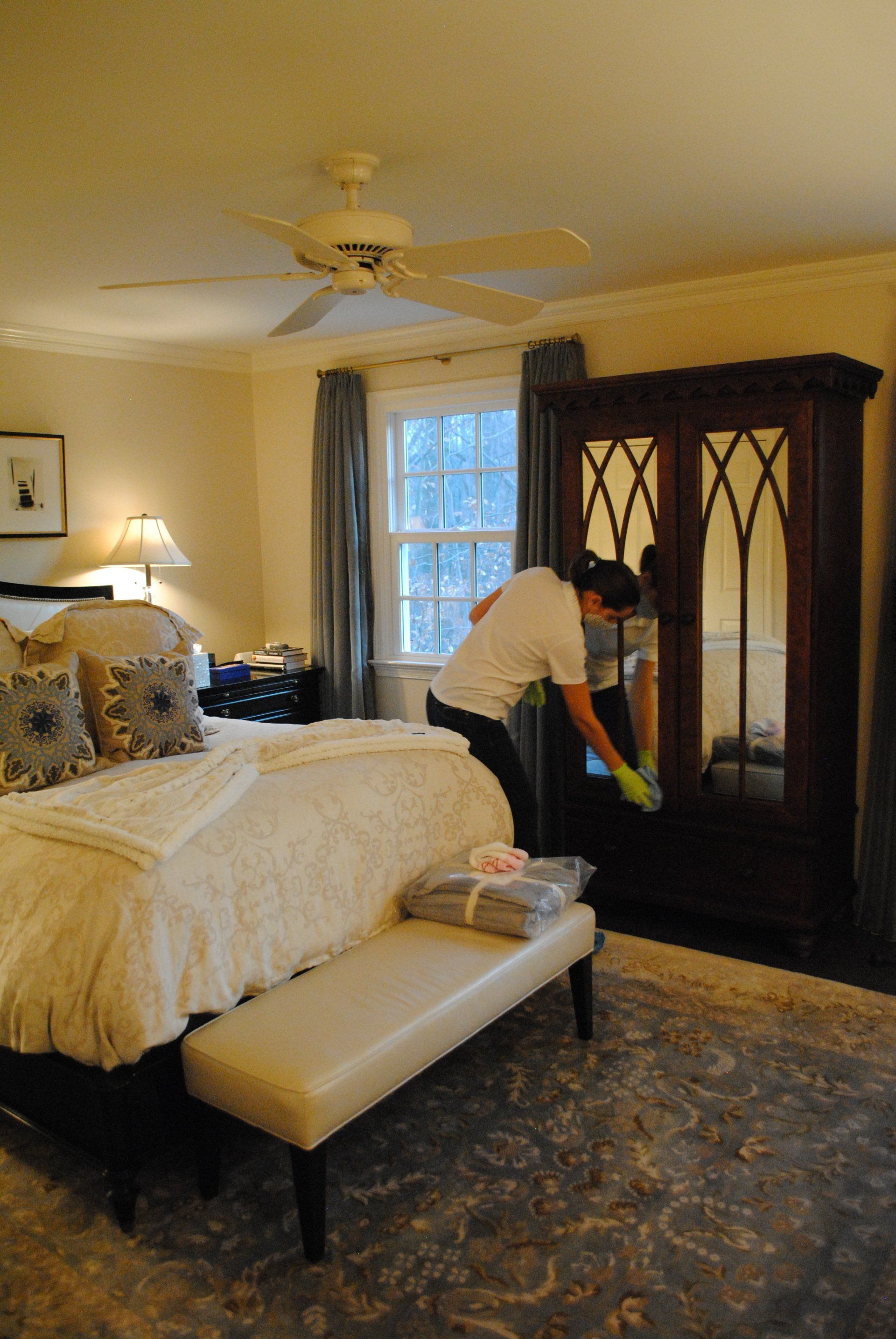 Person making a bed in a cozy bedroom with a dark wood armoire and window.