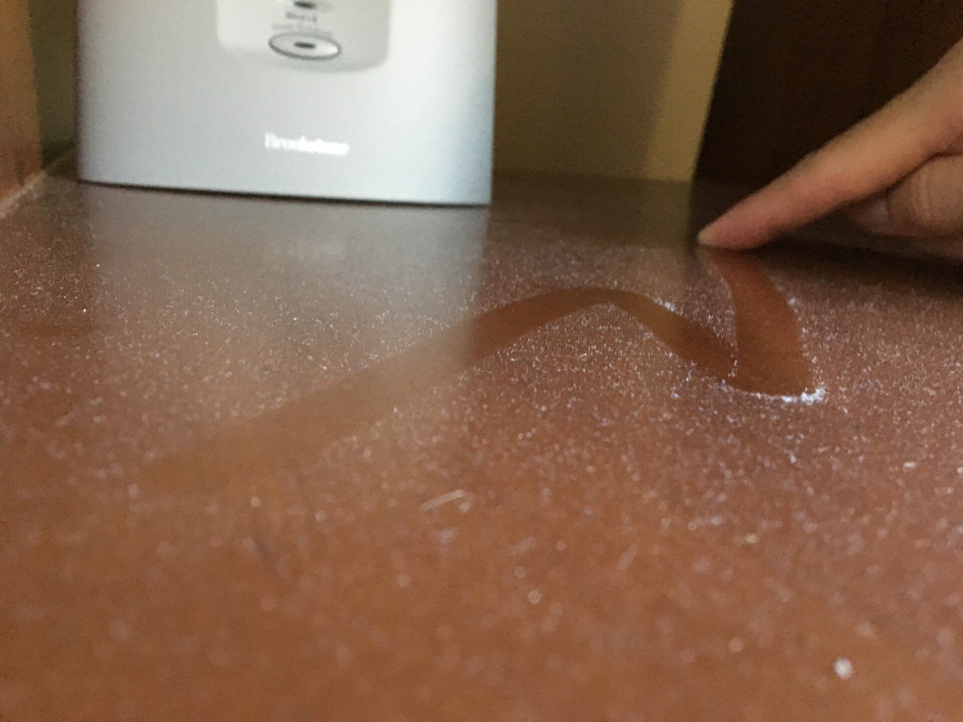 A hand reaches toward a small white appliance with a curved clear lid on a brown tabletop.