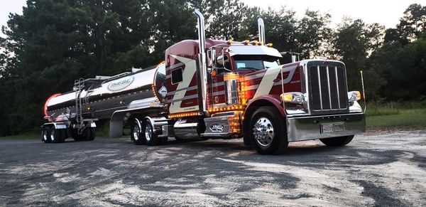 A decorated maroon semi-truck with a tanker trailer parked on gravel in front of a wooded area.