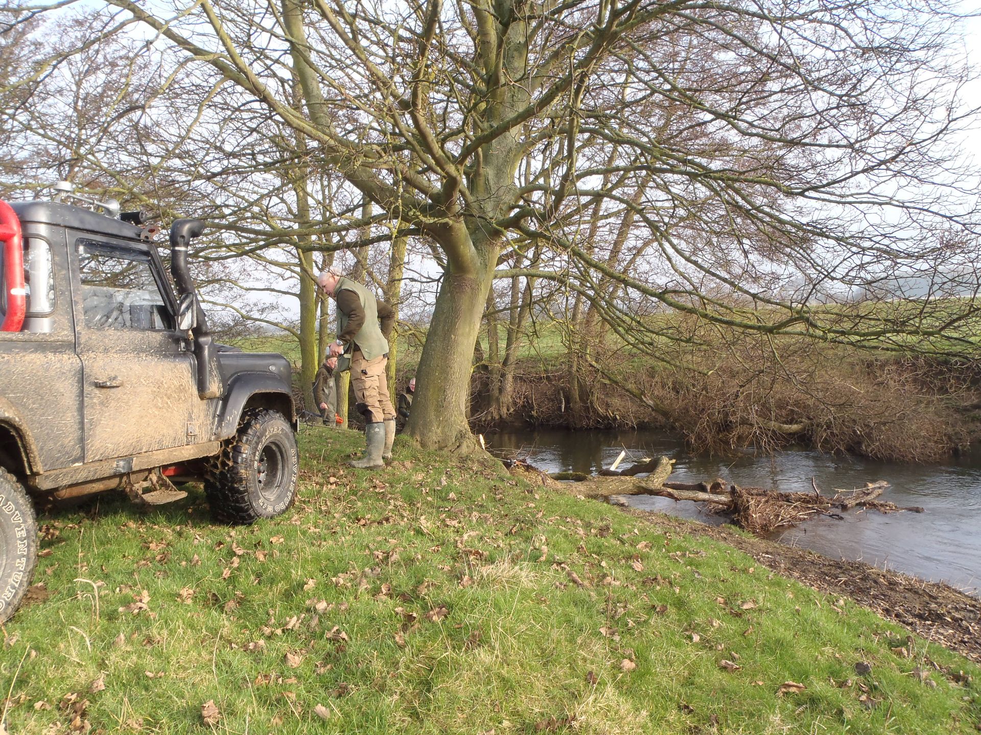 A man is standing next to a muddy jeep near a river.