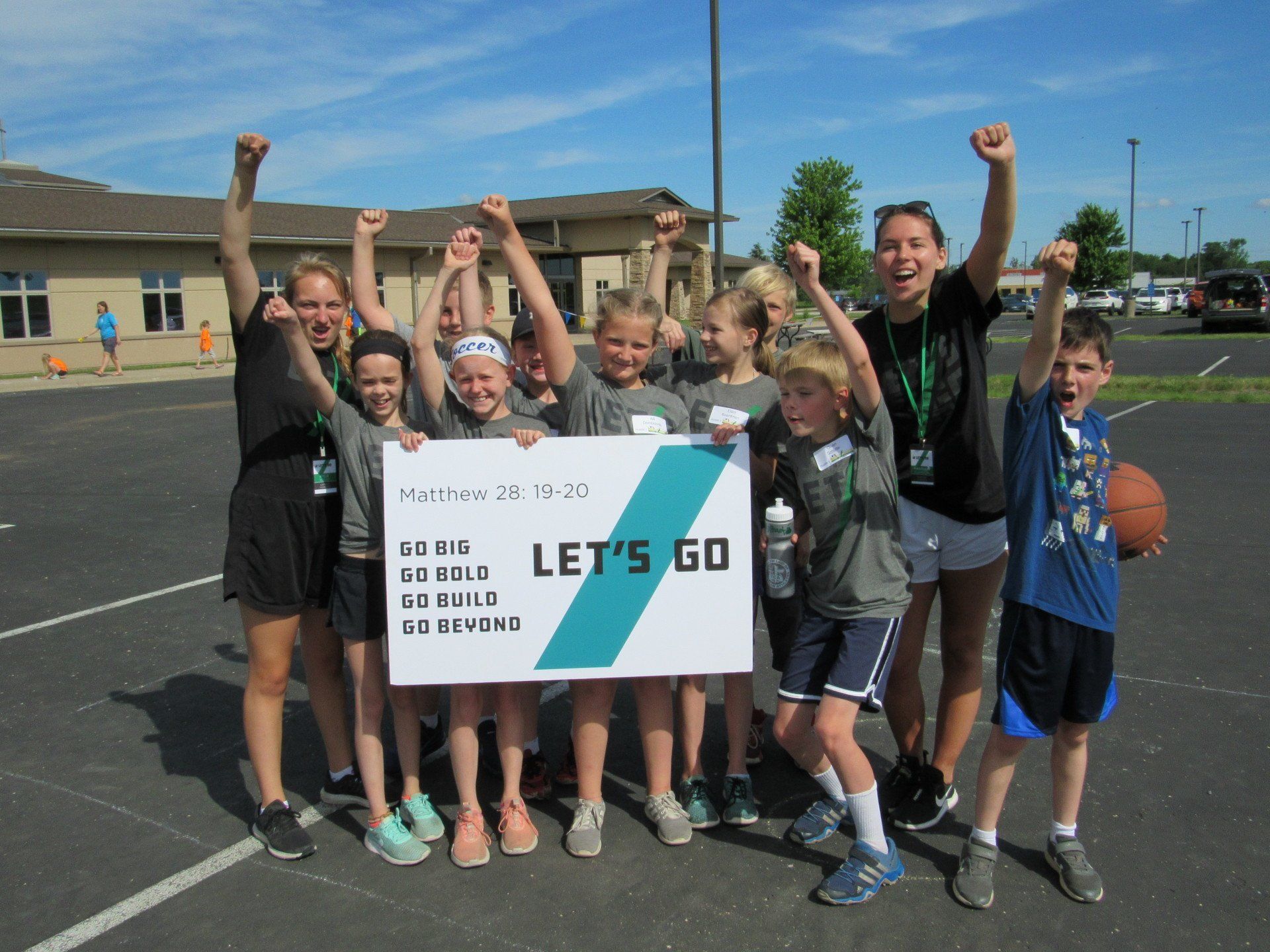 Young athletes with high school leader cheering with arms in the air holding camp sign that says 'Let's Go'