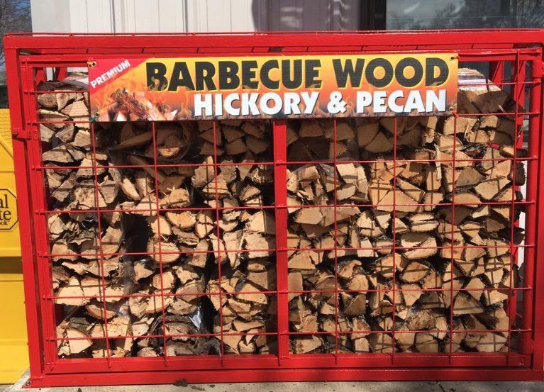 Red metal cage filled with split hickory and pecan barbecue wood. Sign reads