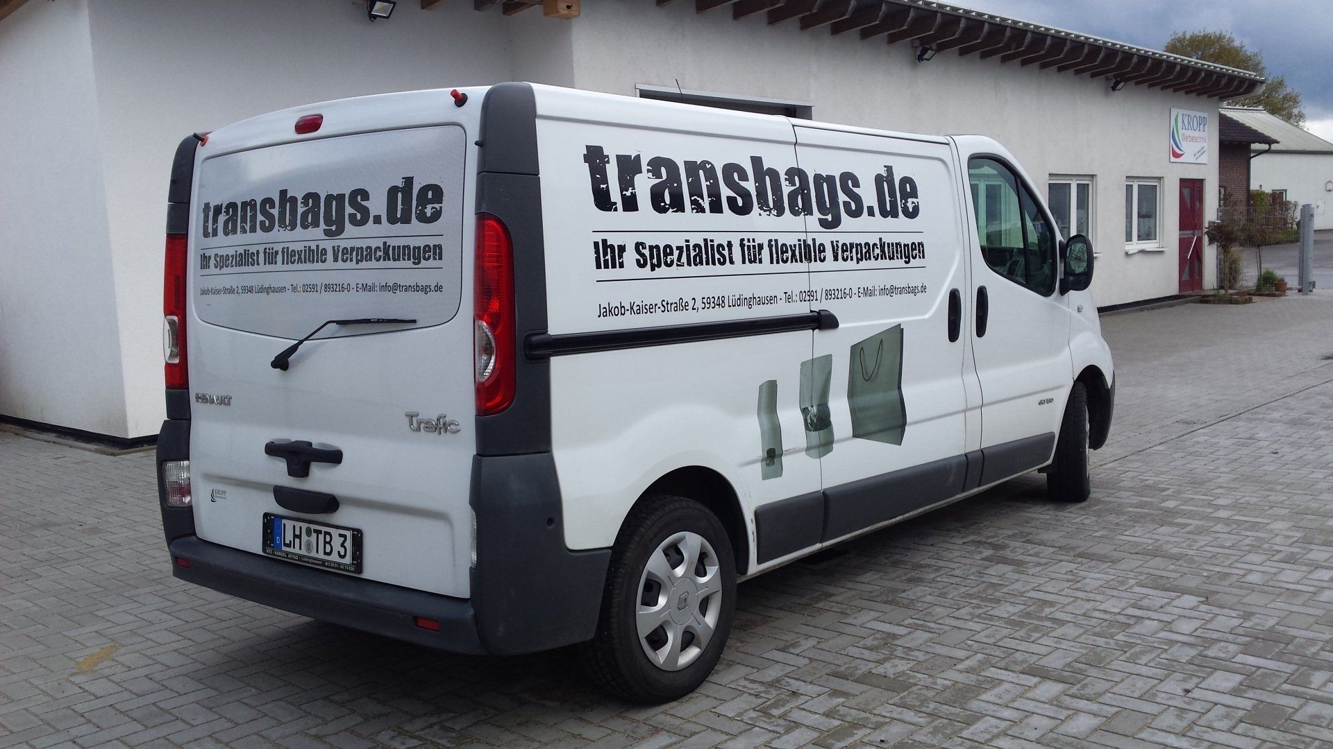 A white van is parked in front of a building that says transbags.de