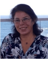 Uma mulher usando óculos e uma camisa florida está sorrindo em frente ao oceano.
