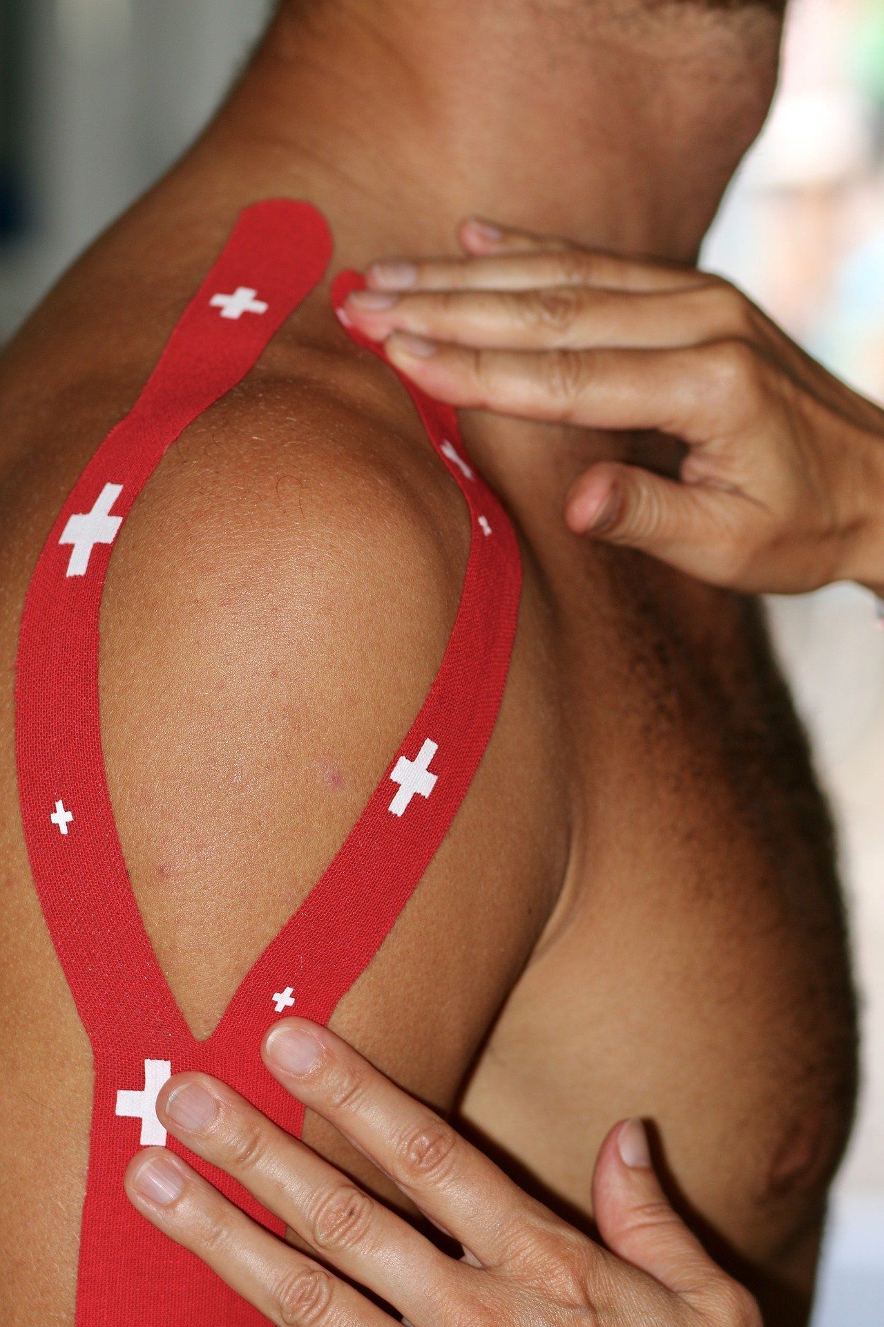 De schouder van een persoon is beplakt met rode kinesiotape; de handen van een andere persoon raken de tape aan.