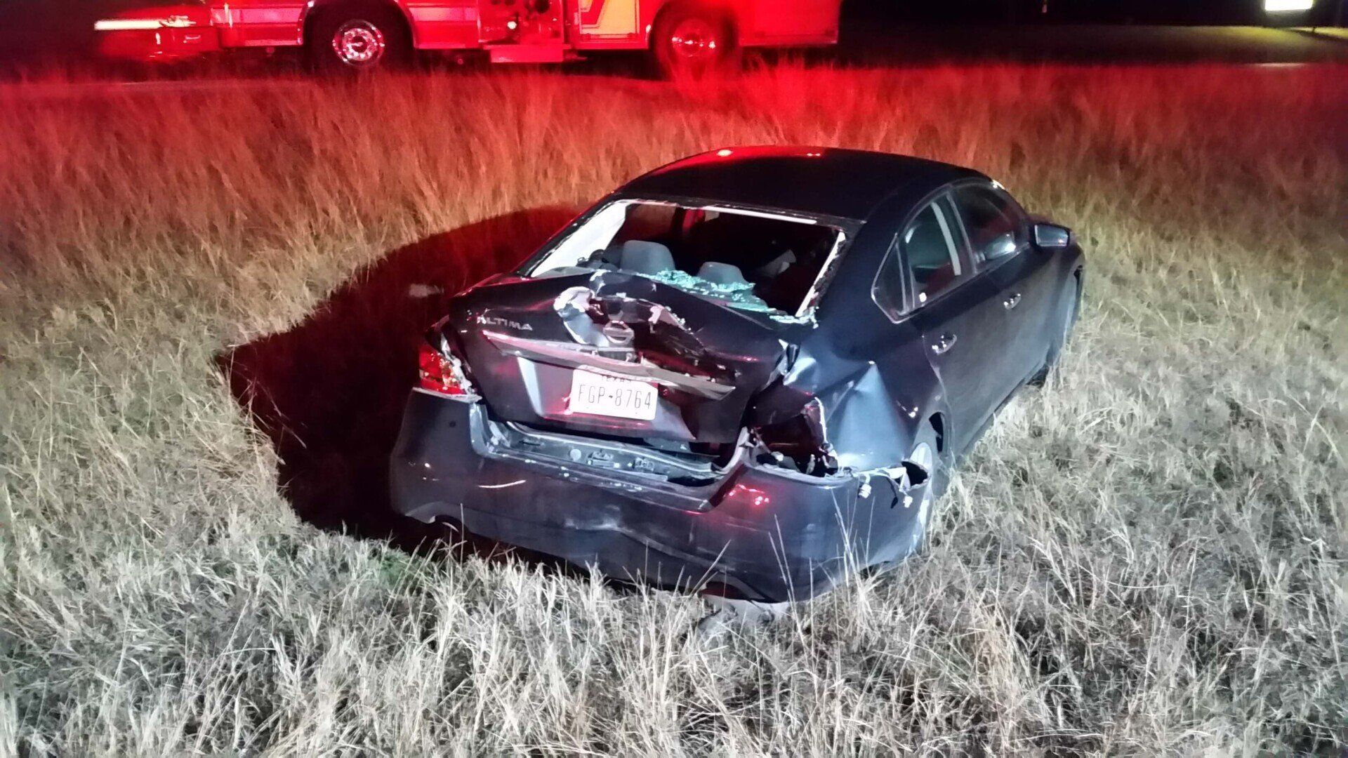 A car with a broken windshield is sitting in the grass next to a fire truck.