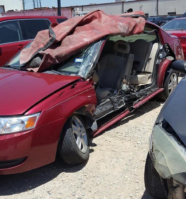 A red car with the hood up is parked in a gravel lot
