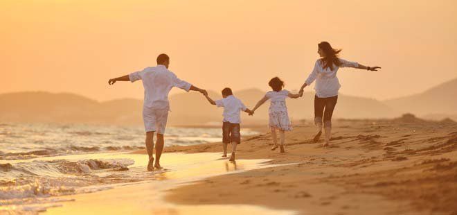 A family of four walking on a beach at sunset, holding hands with arms outstretched.