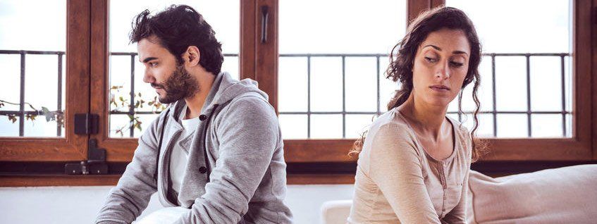 Couple sitting apart, looking down. They appear sad and in distress indoors, near a window.