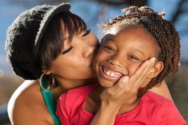 Woman kissing child on the cheek; both smiling outdoors.