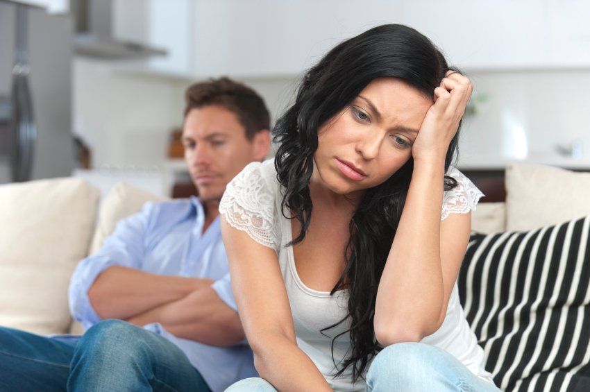 Woman looking sad, hand on head, seated on couch near man with arms crossed, in a living room.