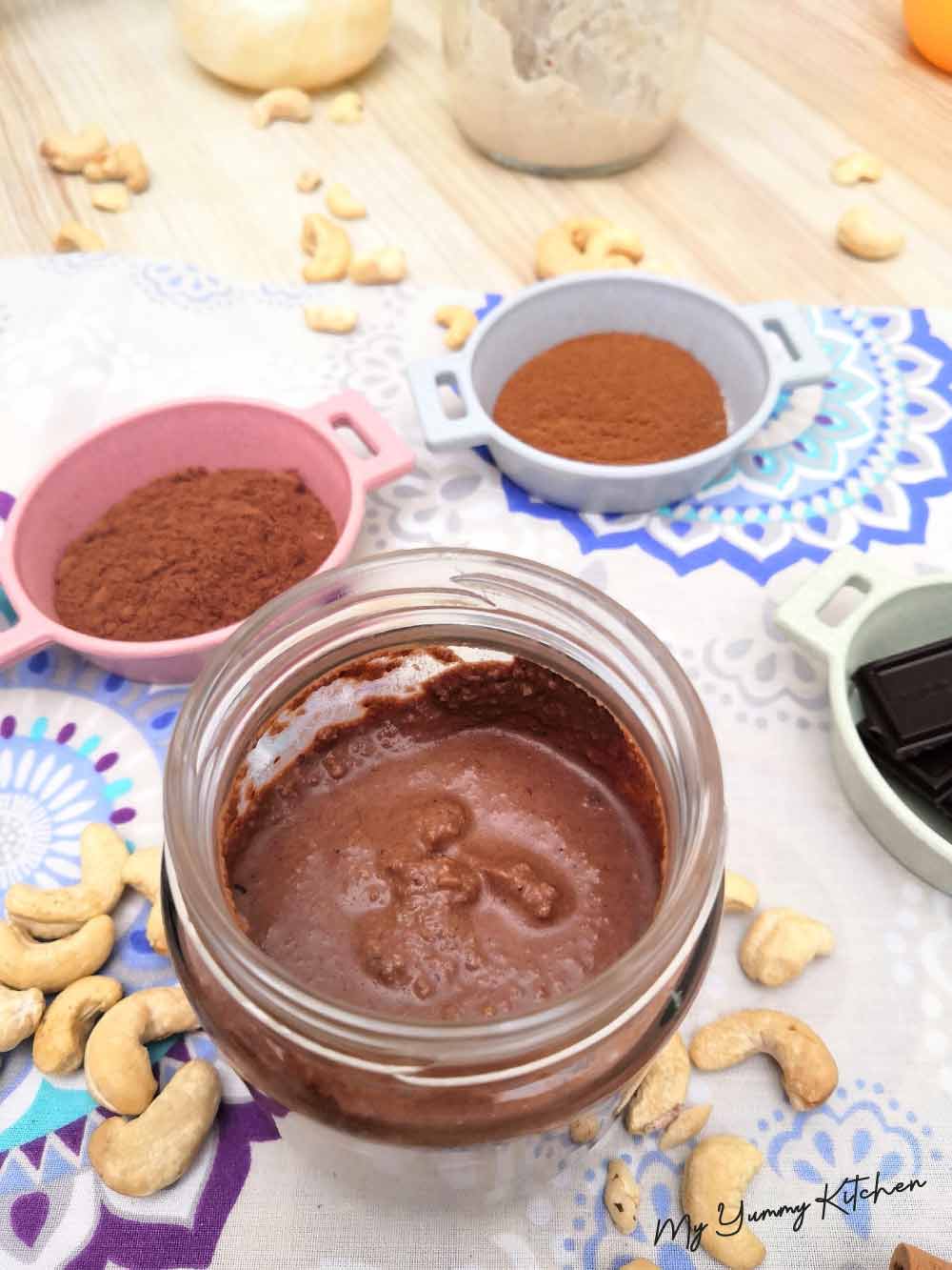 Un pot de pâte à tartiner au chocolat est entouré de tasses à mesurer et de noix de cajou.
