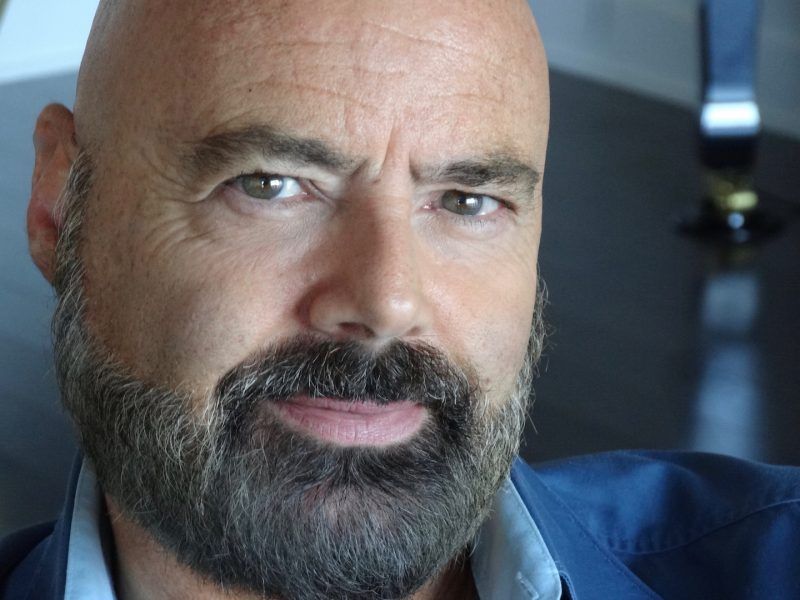 Un homme chauve avec barbe et moustache regarde la caméra.