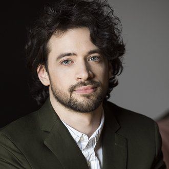 Un homme aux cheveux bouclés et à la barbe porte un costume vert et une chemise blanche.