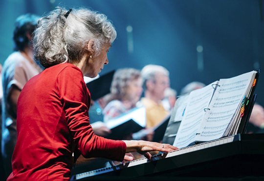 Une femme en chemise rouge joue du piano devant une chorale.